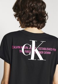 Persona con cabello corto y rizado que lleva una camiseta negra de Calvin Klein Jeans con texto de marca en blanco y rosa en la parte posterior.