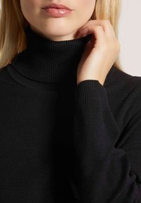Vrouw die de kraag van een zwart geribbeld coltrui aanpast, waarbij lippen, kin, hand en een deel van blond haar te zien zijn.