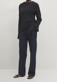 Neulemustat paita pitkillä hihoilla ja pyöreällä pääntiellä, paritettu tummansinisten leveälahkeisten housujen ja mustien hihnallisten sandaalien kanssa.