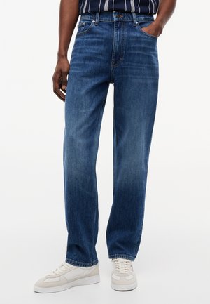 Άνδρας που φορά άνετα μπλε τζιν και μπεζ αθλητικά παπούτσια, με το ένα χέρι στην τσέπη και ριγέ πουκάμισο μέσα στο παντελόνι.