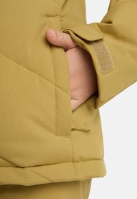 Detail einer gelben, gepolsterten Jacke mit Seitentasche und Klettverschluss am Ärmel, weicher, matter Textur und robuster Naht.