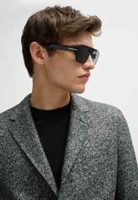 Occhiali da sole neri con design geometrico, lenti scure e aste sottili con accenti argentati, abbinati a un blazer grigio strutturato.