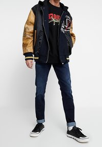 Chaqueta varsity negra y marrón con detalles gráficos, sudadera negra, jeans de mezclilla oscuros y zapatillas negras con acentos blancos.