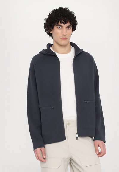 Hanorac cu fermoar de culoare bleumarin închis, cu glugă, fabricat dintr-un material moale, cu buzunare frontale cu fermoar, asociat cu un tricou alb și pantaloni cargo bej deschis.