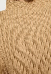 Jersey de punto con cuello alto en color beige claro. La tela presenta un patrón texturizado con costuras visibles.