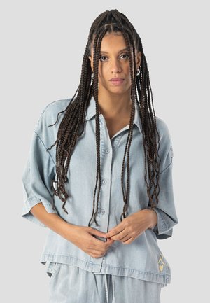 Kvinde med lange fletninger iført lys denimskjorte og matchende bukser, stående mod en ensartet lysegrå baggrund.