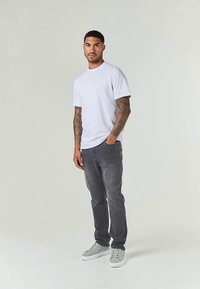 Homem com os braços tatuados, vestindo uma t-shirt branca simples, jeans cinza escuro e tênis cinza claro, de pé contra um fundo claro e simples.