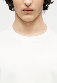 Jauna persona ar gaišu ādu un tumšiem, cirtainiem matiem, valkājot vienkāršu baltu T-kreklu ar apaļu kaklu, tuvplānā no zoda līdz augšējiem krūtīm.