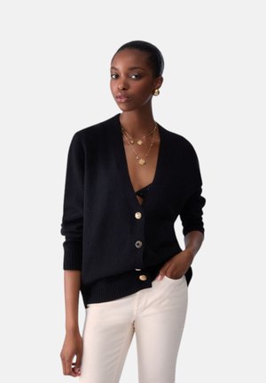 Donna che indossa un cardigan nero con bottoni, pantaloni bianchi, orecchini dorati e collane dorate a strati, con una mano in tasca.