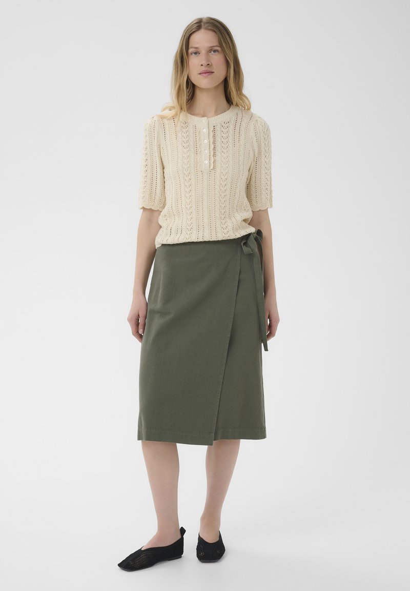 Femme portant un pull court crème en maille, une jupe portefeuille midi vert olive nouée à la taille et des chaussures plates noires, debout sur un fond blanc.
