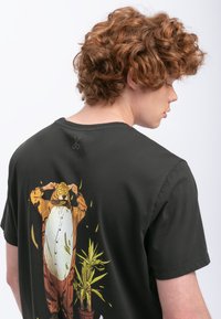 Zwarte t-shirt van katoen, met een kleurrafische afbeelding op de achterkant van een karakter in een tijger onesie met planten eronder.