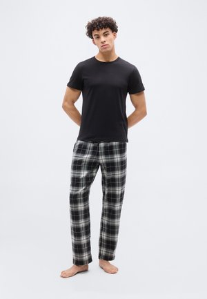 Unge mand stående barfodet, iført en ensfarvet sort t-shirt og sort-hvide ternede pyjamasbukser mod en ensfarvet lys baggrund.