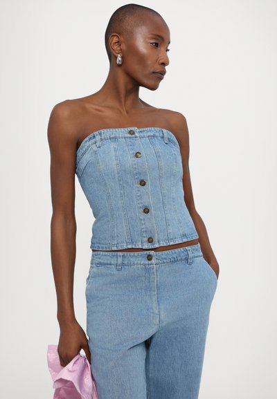 Jeans uten skuldre topp med en tettsittende design, lys blå farge, og fem knapper foran. Modellen holder en rosa stoffbit i den ene hånden.