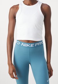 Persona atlética vestida con un top corto sin mangas blanco y leggings Nike Pro azules, de pie con los brazos relajados a los lados.