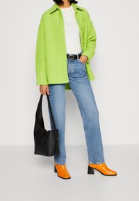 Neon-grünes, übergroßes Hemd als Jacke, enges weißes Oberteil, hellblaue Jeans, schwarze Handtasche und glänzende orangefarbene Stiefeletten mit Blockabsatz.