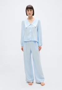 Γυναίκα που στέκεται ξυπόλητη, φοράει ανοιχτό μπλε χαλαρή πιτζάμα με μεγάλο γιακά και πουκάμισο με κουμπιά και μακριά μανίκια, μπροστά σε απλό φόντο.