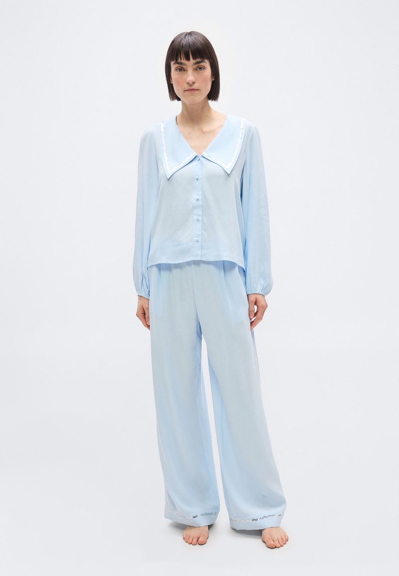 Γυναίκα που στέκεται ξυπόλητη, φοράει ανοιχτό μπλε χαλαρή πιτζάμα με μεγάλο γιακά και πουκάμισο με κουμπιά και μακριά μανίκια, μπροστά σε απλό φόντο.