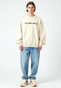 Felpa oversize beige con testo nero, abbinata a jeans baggy azzurri chiari e sneaker bianchi chunky con dettagli rossi e blu.