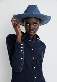 Jasnoniebieska koszula denimowa z guzikami w kolorze białym i szyciem w stylu zachodnim, zestawiona z szerokim kapeluszem denimowym w odcieniu niebieskim z paskowanymi akcentami.