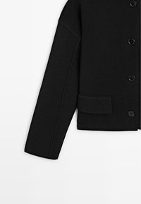 Manica di giacca nera in maglia con chiusura a bottoni e una tasca frontale rettangolare su sfondo bianco.