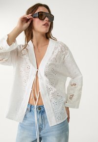 White embroidered blouse with a deep V-neck and adjustable tie closure. Loose fit, floral cut-outs, and wide sleeves. Worn with blue jeans.