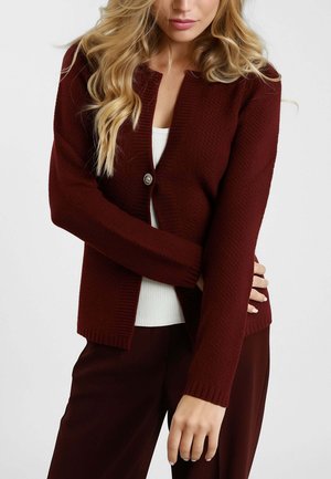 Donna che indossa un cardigan testurizzato bordeaux con un bottone su una maglietta bianca e pantaloni bordeaux abbinati, in posa con le braccia incrociate.