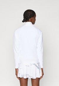 Balta ilga rankovėmis palaidinė su aukštu krageliu, lygaus tekstūro ir prigludusia fasonu, derinama su šviesia, taškinio rašto sijonu.