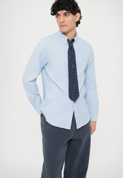 Vaaleansininen puuvillainen nappipaita, jossa kaulus, yhdistetty tummansiniseen solmioon, jossa on painettu kuvio. Harmaat teksturoidut housut viimeistelevät asun.