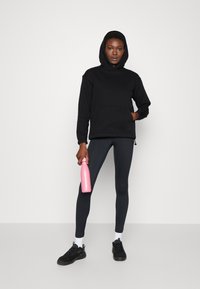 Felpa con cappuccio nera, leggings neri, sneakers neri, tiene in mano una bottiglia d'acqua rosa. Design minimalista, vestibilità casual, texture liscia.