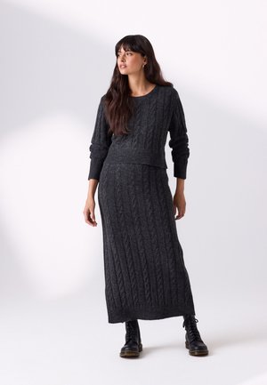 Femme aux longs cheveux foncés portant un pull en torsades gris foncé et une longue jupe assortie, associée à des bottes noires à lacets, debout.