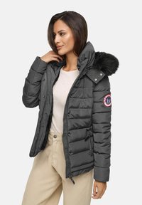 Vrouw in een donkergrijze gewatteerde jas met een met bont afgewerkte capuchon, die de kraag vasthoudt, in combinatie met beige broeken en een licht shirt.