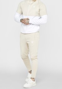 Beige mikina na zip s bílými rukávy a odpovídajícími béžovými tepláky. Obsahuje mírný lesk a výšivku loga na jak mikině, tak na kalhotách. Bílé tenisky.