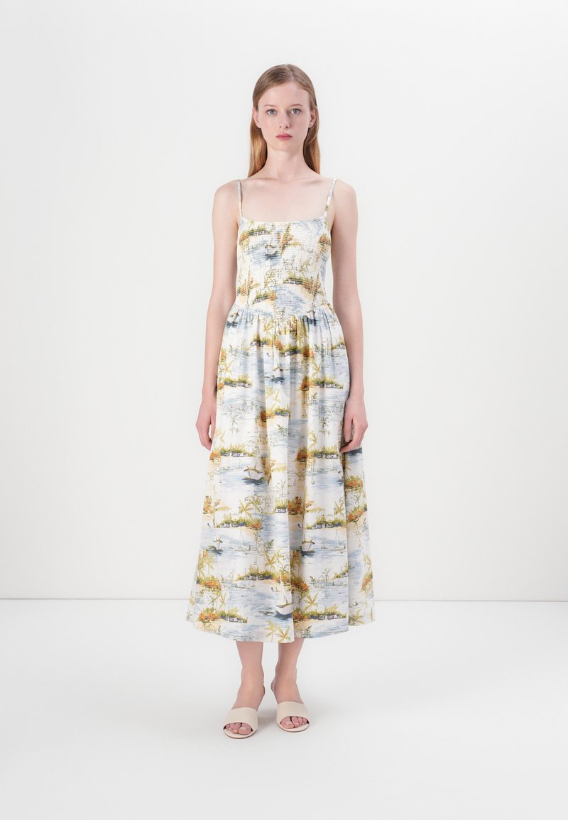 Robe maxi sans manches à motifs floraux en bleu clair avec des accents jaunes et verts. Tissu fluide avec taille froncée. Sandales blanches.