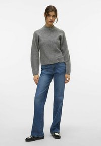 Suéter gris de punto con cierre de cremallera, cuello alto y mangas acanaladas. Combinado con pantalones vaqueros de campana de color azul claro y zapatos negros.