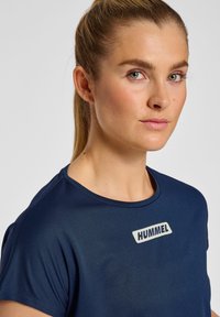 Mörkblå kortärmad sportskjorta med en slät yta och en framträdande "HUMMEL"-logotyp i vitt på bröstet.