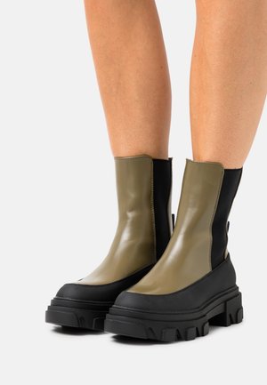 Botas tobilleras robustas con parte superior en oliva mate y paneles elásticos negros, que cuentan con una suela gruesa de goma negra con un patrón de banda de rodadura texturizado.