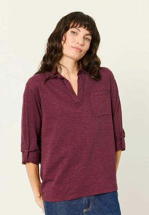 Woman with wavy brown hair wearing a long-sleeve, buttoned-up maroon shirt and blue jeans, standing against a plain light background.