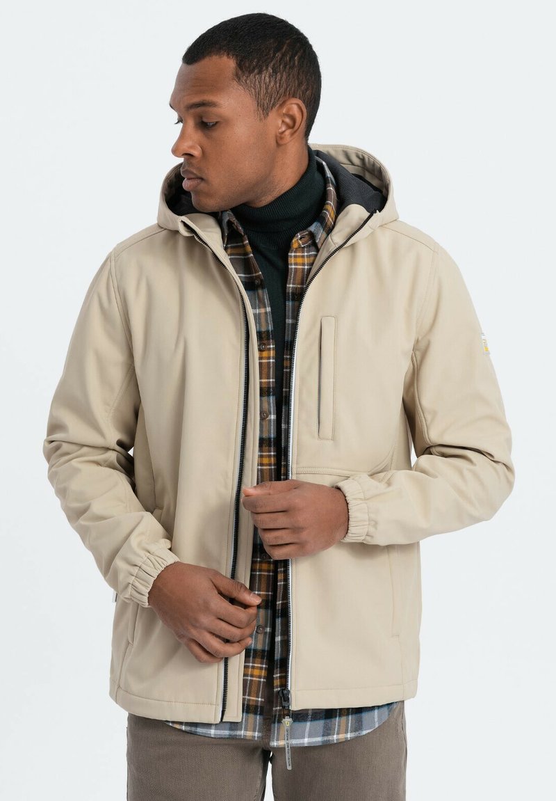 Beige wasserdichte Jacke mit Kapuze, elastischen Bündchen, Frontreißverschluss und Seitentaschen über einem dunkel karierten Flanellhemd. Glatte Textur.