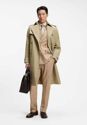 Mand iført beige trenchcoat over tan jakkesæt med mønstret slips, der holder en mørk brun lædermappe og står mod hvid baggrund.