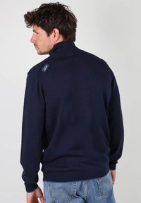 Pull en maille bleu marine avec un col montant, des poignets et un ourlet côtelés, arborant un logo cousu sur l'épaule arrière. Texture douce, coupe décontractée.