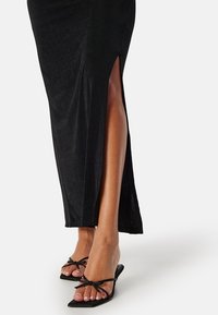 Zwarte maxi-jurk met een zijsplit, met een zachte textuur. Gecombineerd met zwarte platte sandalen met dunne, geknoopte banden.