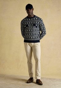 Marineblauer Pullover mit weißen Motiven, versehen mit dem Text "BAA HUMBUG." Kombiniert mit beigen Hosen und braunen Schuhen. Eine schwarze Mütze rundet den Look ab.