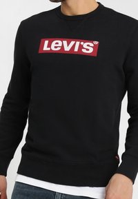 Homme portant un sweat-shirt noir Levi's avec un logo rouge et blanc sur la poitrine, superposé à une chemise blanche et un jean bleu.
