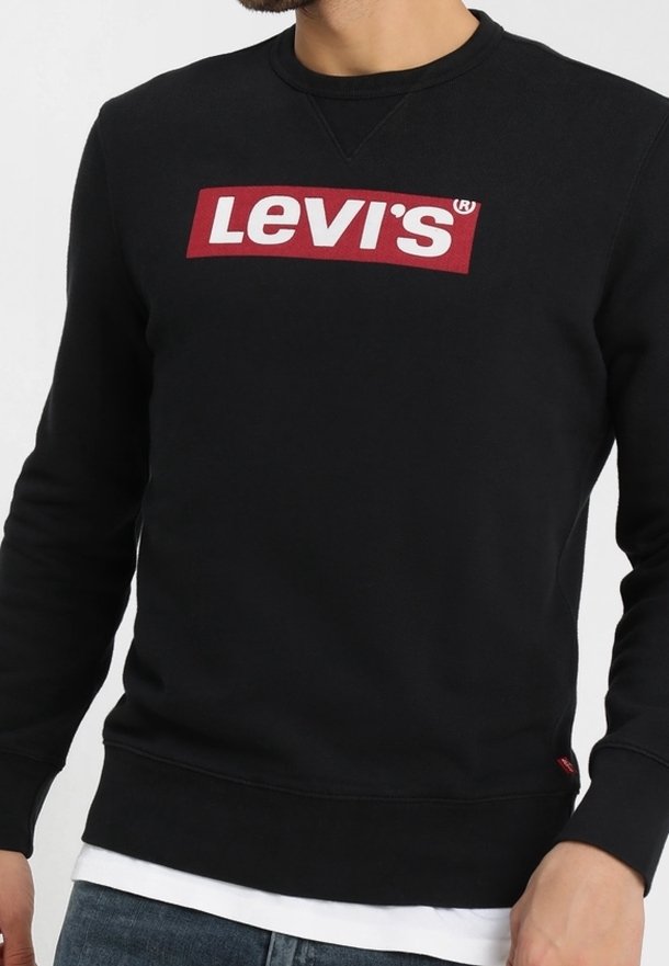 Homme portant un sweat-shirt noir Levi's avec un logo rouge et blanc sur la poitrine, superposé à une chemise blanche et un jean bleu.