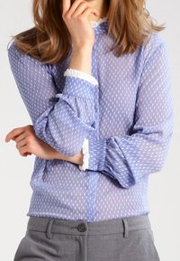 Blouse légère bleu clair en voile avec des pois blancs irréguliers, col volanté et fermeture à boutons à l'avant, dotée de manches à boutons et d'une coupe flexible.