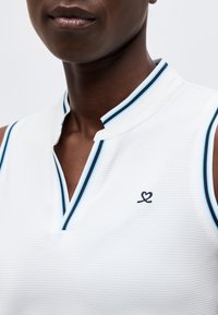 Κοντινό πλάνο ενός ατόμου που φοράει λευκή αμάνικη μπλούζα με μπλε και μαύρη ριγέ επένδυση και μικρό κεντημένο λογότυπο καρδιάς κοντά στον γιακά.