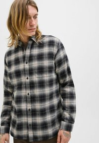 Camisa de franela a cuadros en negro, gris y crema, con cierre de botones, mangas largas y un bolsillo de parche en el pecho.