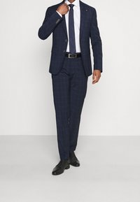 Completo a quadri navy con vestibilità su misura, giacca monopetto e pantaloni con tasche piatte. Abbinato a una camicia bianca e una cravatta scura, indossato con scarpe nere.