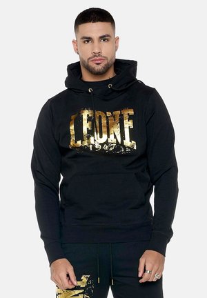 Mann trägt schwarzen Hoodie mit goldener Aufschrift „LEONE 1947“ und passende schwarze Hose mit goldenem Design, steht vor weißem Hintergrund.