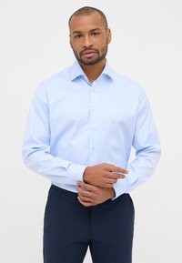 Camicia azzurra chiara con colletto, caratterizzata da una texture liscia e maniche lunghe. Abbinata a pantaloni scuri. I bottoni sono bianchi e rotondi.